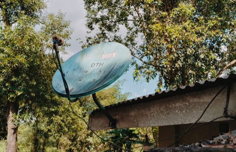 Antena parabólica em um telhado rural, cercada por árvores verdes.